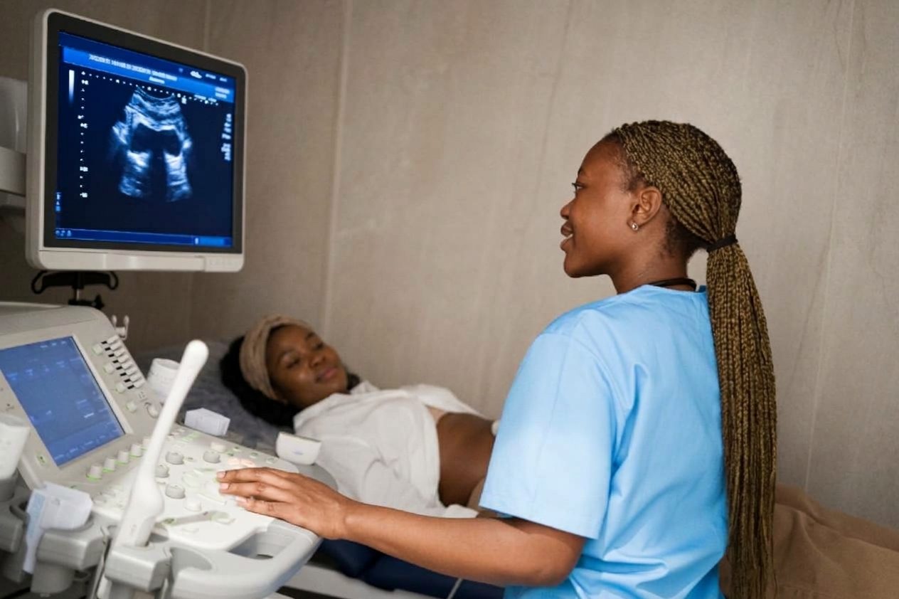 A clinician in blue scrubs operates an ultrasound machine while a patient lies on an examination table