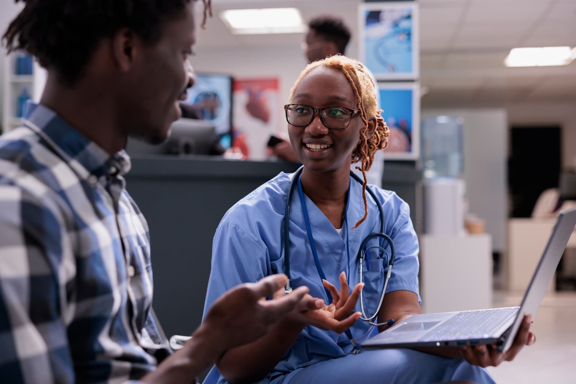 A nurse in blue scrubs holding a laptop talks with a seated patient in a clinic room