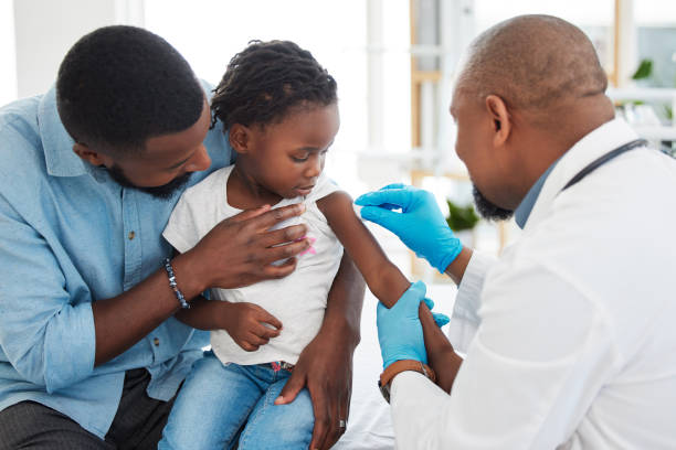 A father sits close beside his young daughter as a clinician in a white coat and blue gloves prepares her arm for a vaccination