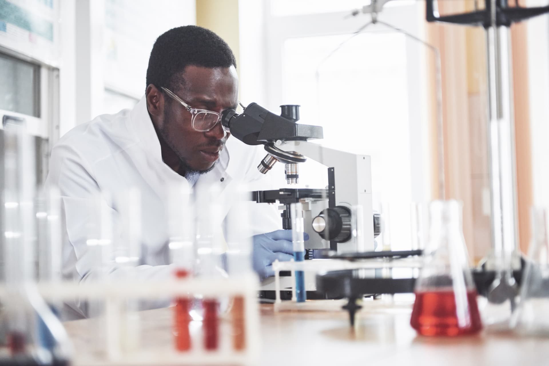 A scientist in a white lab coat, glasses and gloves looks through a microscope in a laboratory filled with glassware
